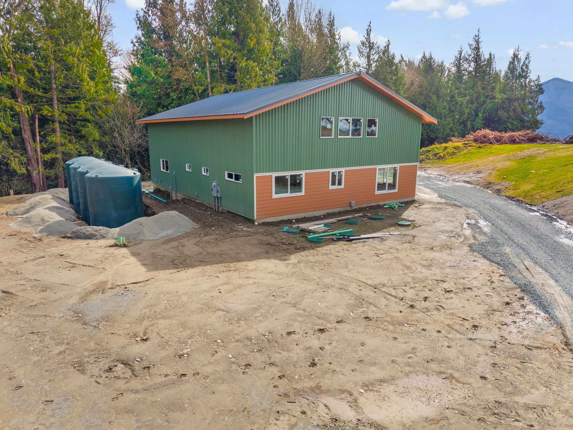 A two-story building with green metal siding and brick foundation sits on a dirt lot next to three large water tanks.