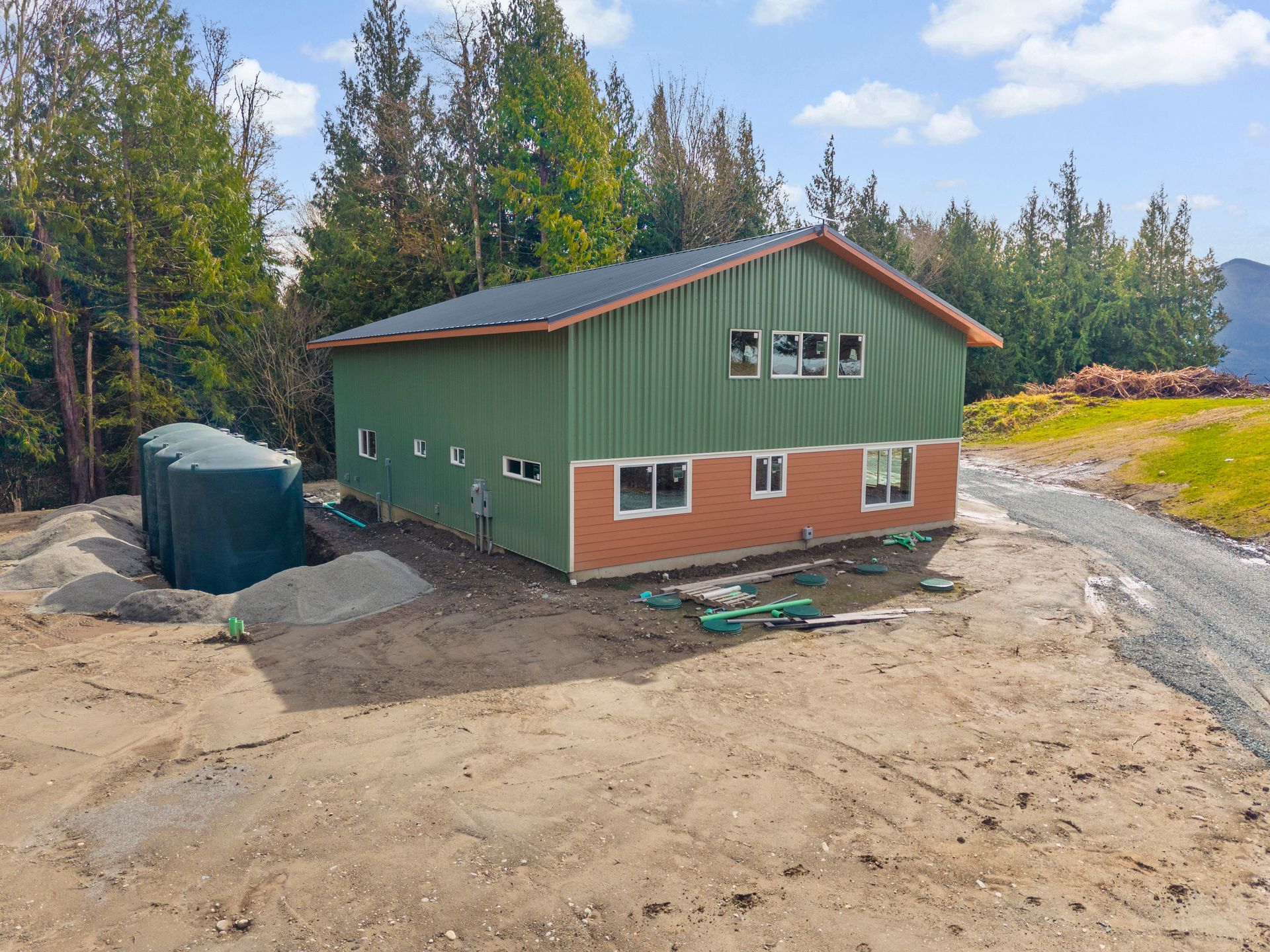 A green building with a brick lower level and black roof, positioned on a gravel lot next to three large water tanks.