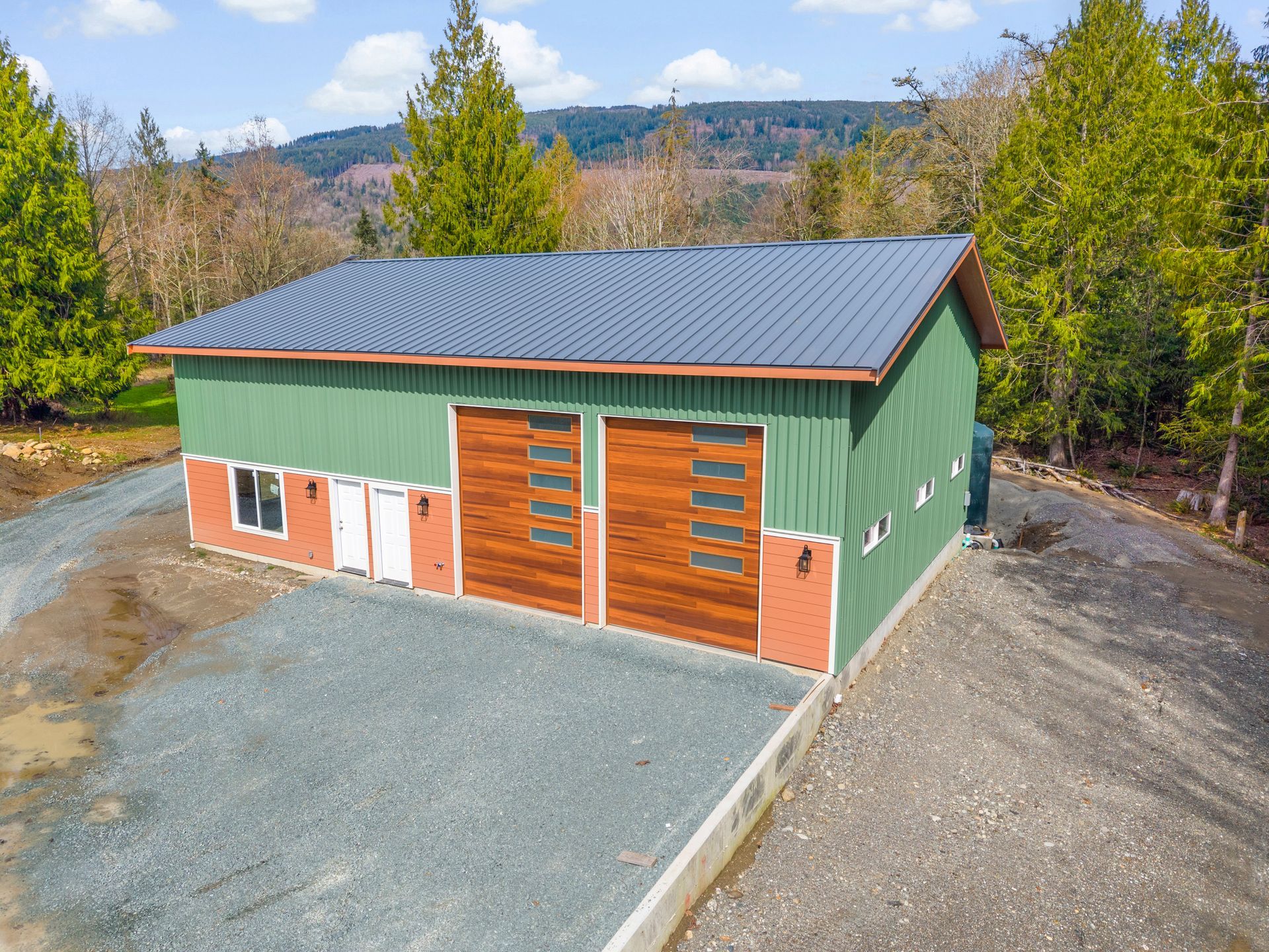 A green metal shop building with wood-look garage doors, a red brick-patterned base, and a grey roof, set in a wooded area.