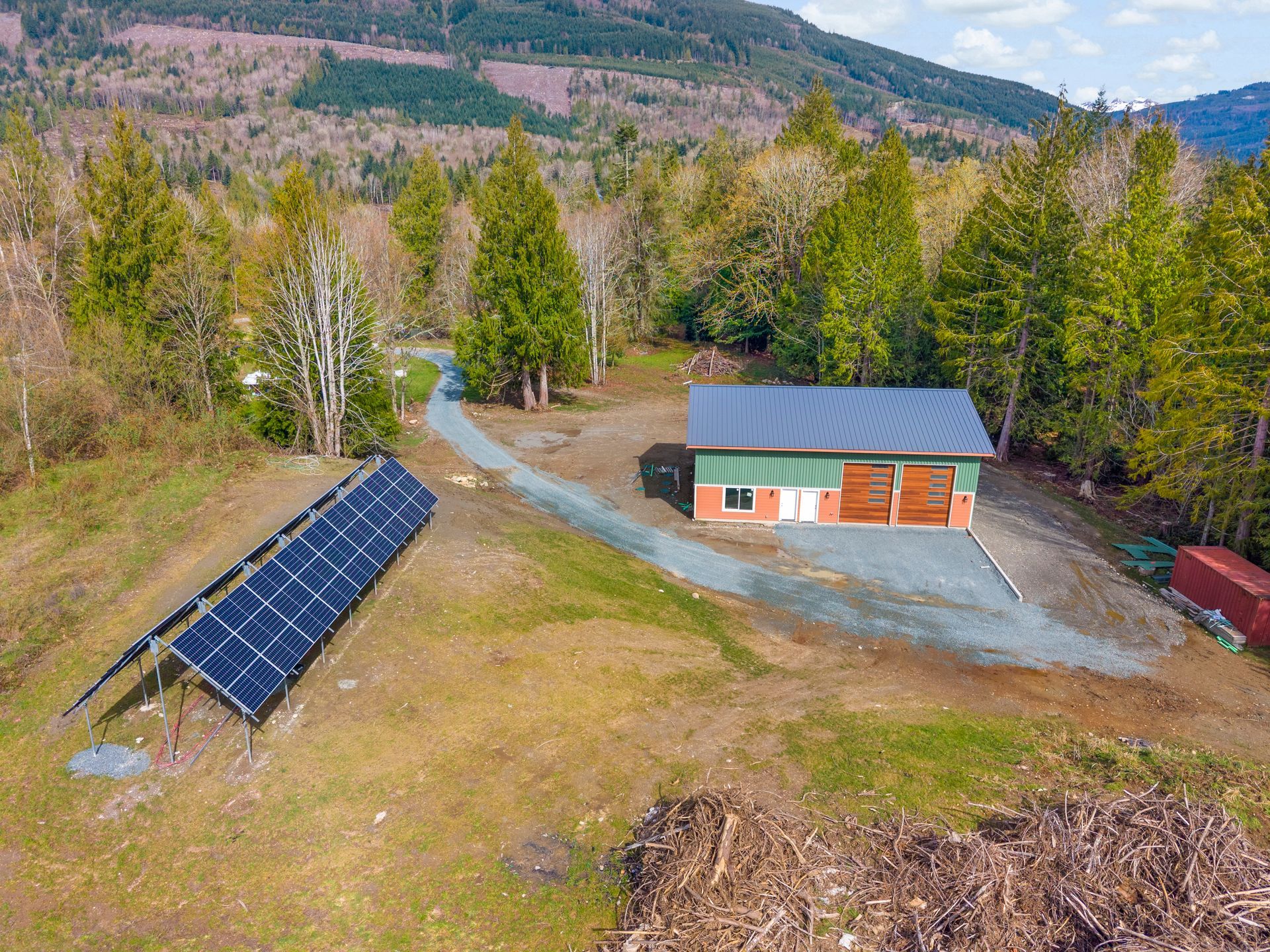 Aerial view of a detached garage with solar panels in a clearing surrounded by woods and a hill.