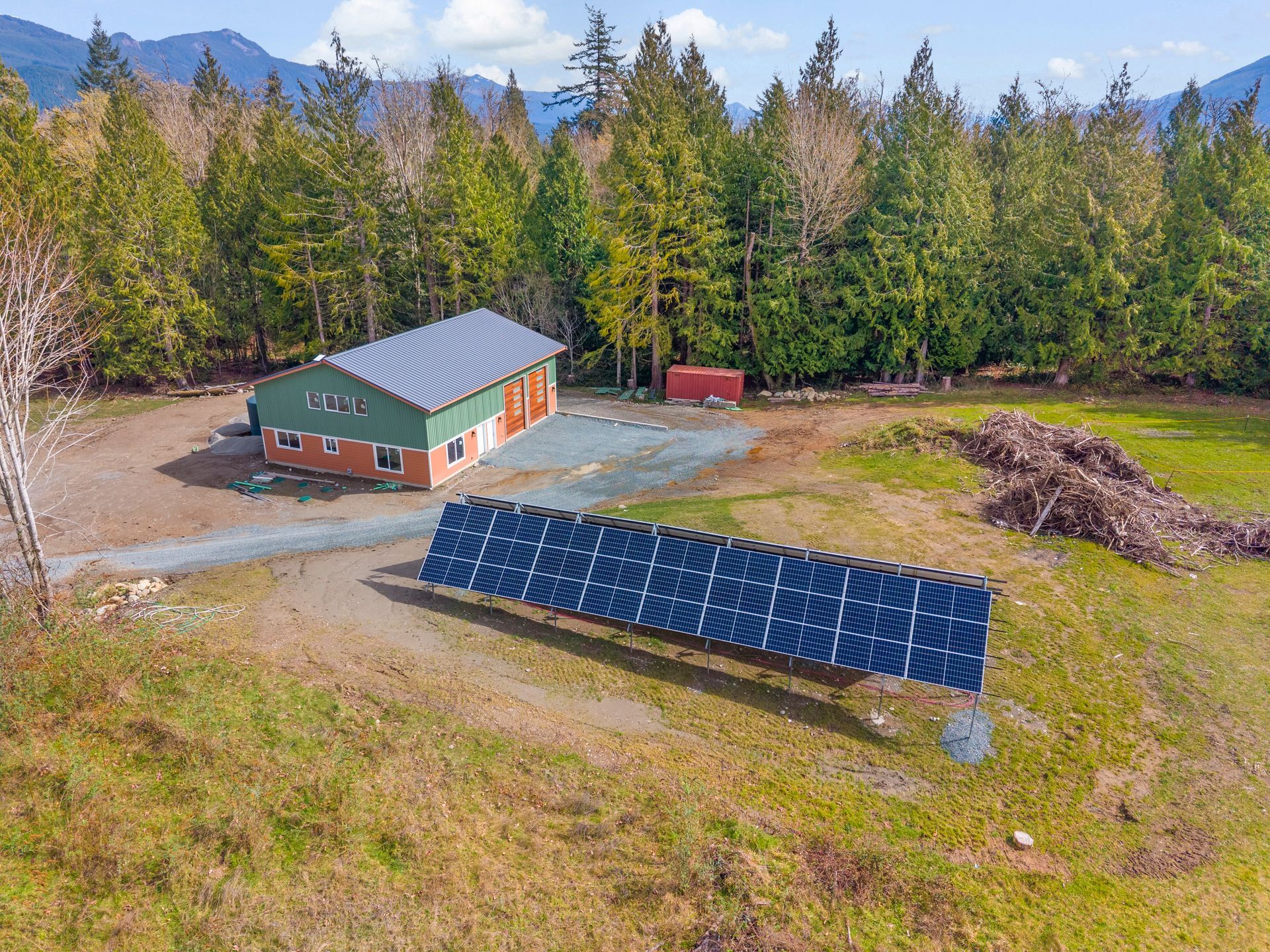 A large ground-mounted solar panel array sits in a grassy field in front of a green and brick building near a forest.