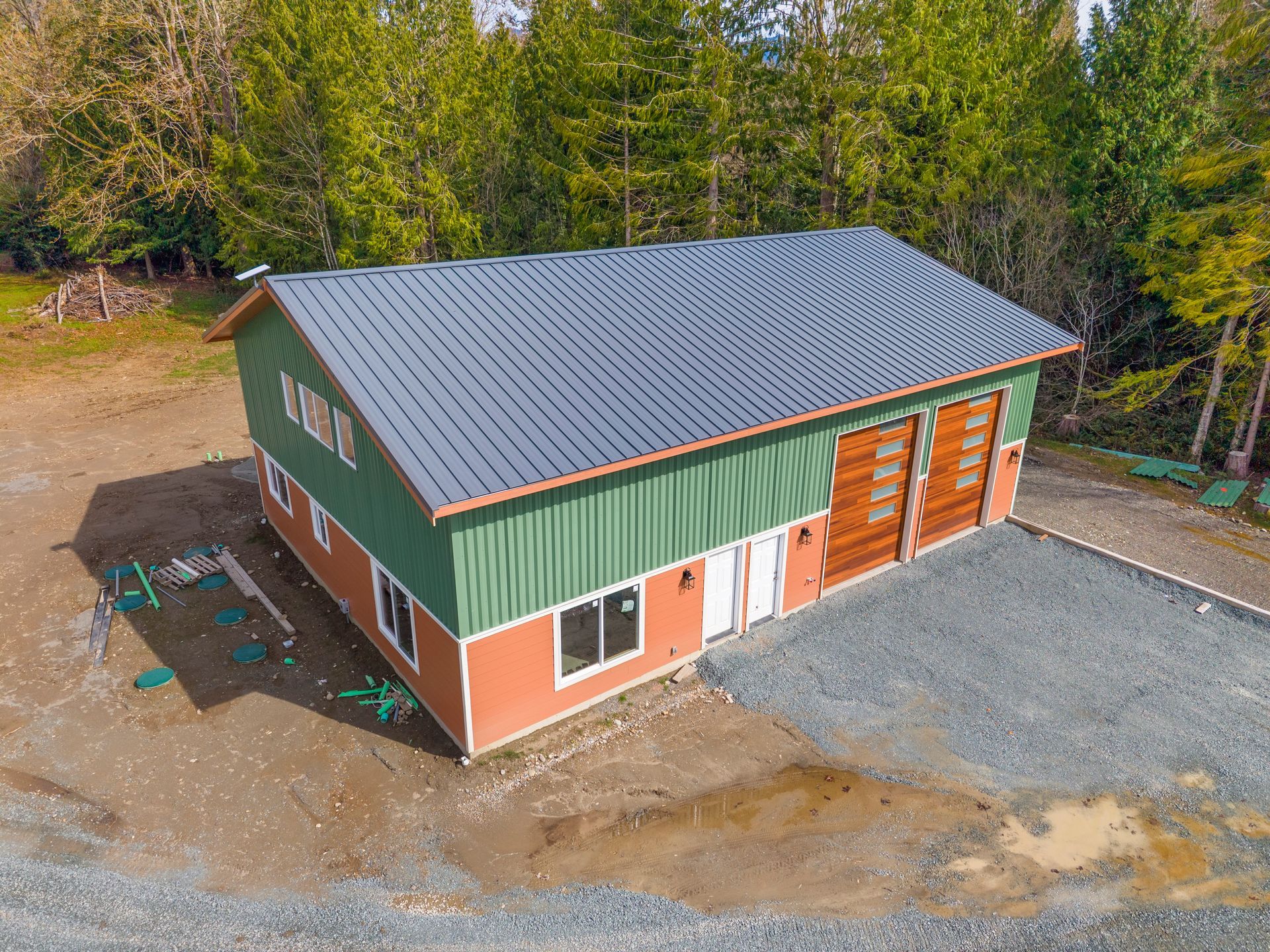 An aerial view of a two-story building with a grey metal roof, green siding, and orange trim, set in a forested area.
