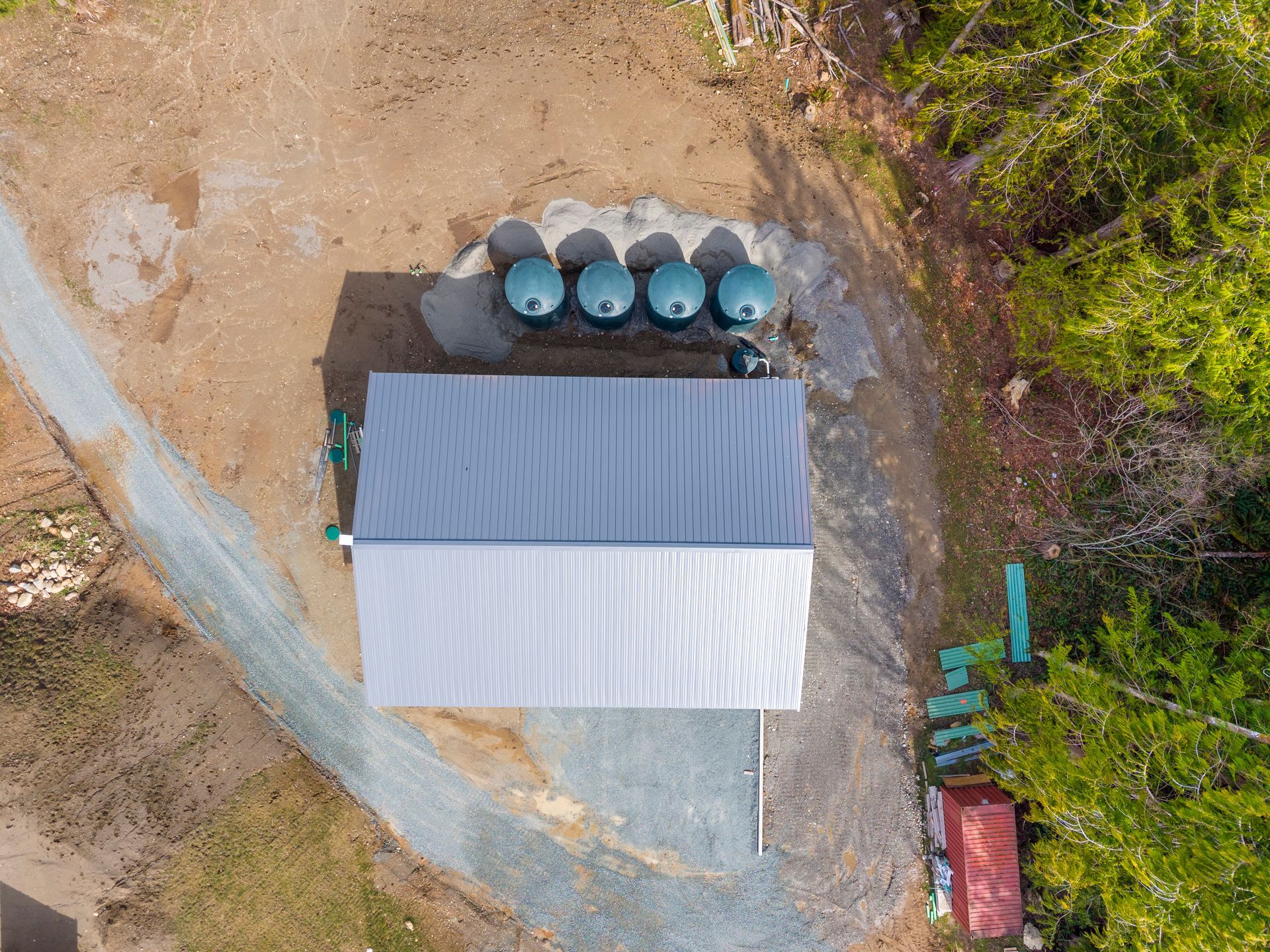 An aerial view of a shed with a silver metal roof, four water tanks nearby, and a gravel driveway in a wooded area.