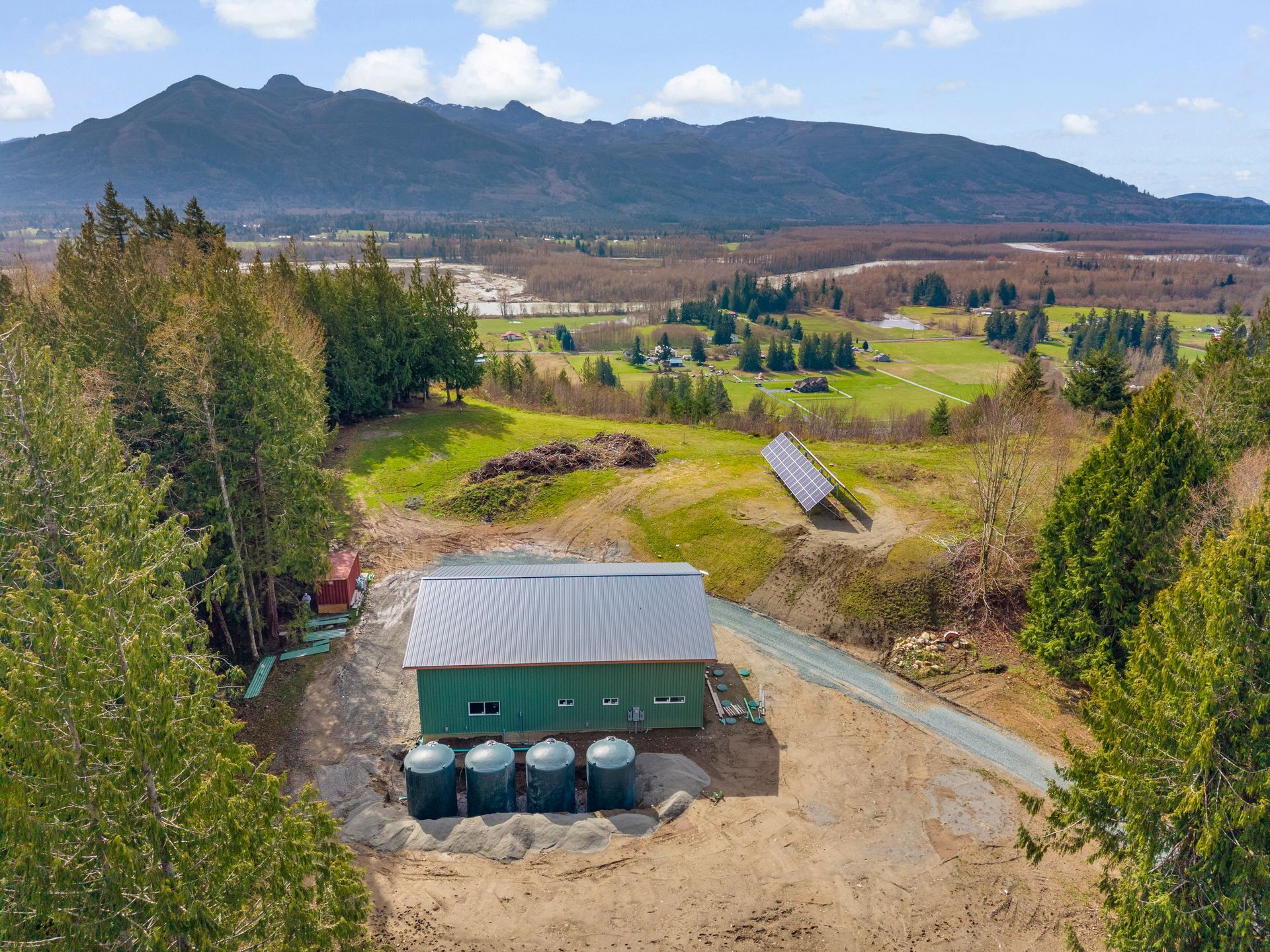 A green building with a gray roof and four water tanks sits in a grassy, rural landscape with mountains in the background.