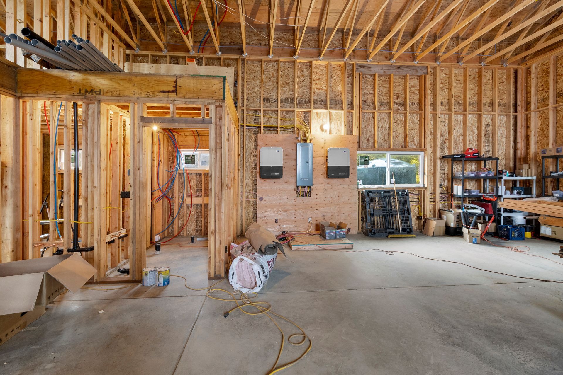 A construction site interior with wooden framing, exposed walls, an electrical panel, and storage shelves on a concrete floor.