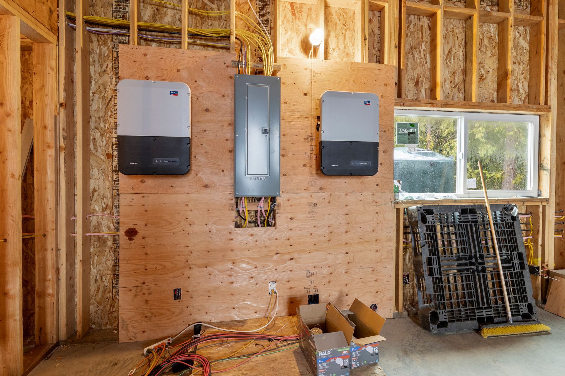 Two solar inverters flank a central electrical panel mounted on a plywood wall in an unfinished construction room.