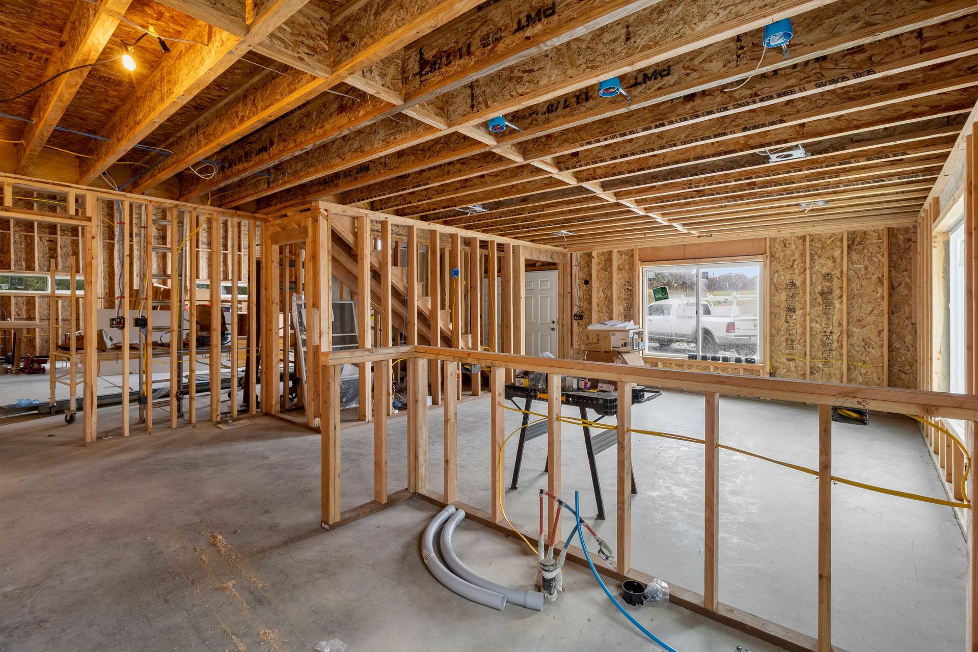 Interior of a room under construction with exposed wooden wall studs, floor joists, concrete flooring, and a large window.