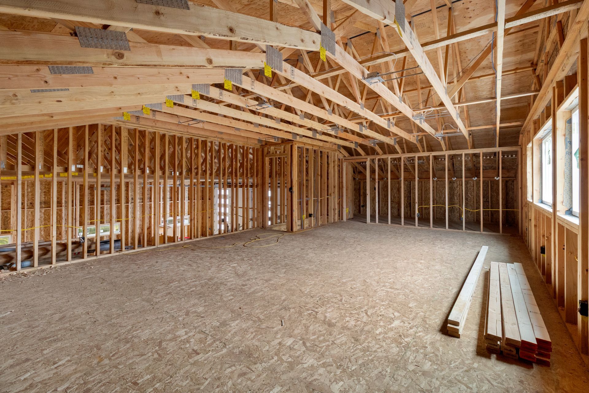 The interior of a building under construction, featuring wooden wall framing and roof trusses over a plywood floor.