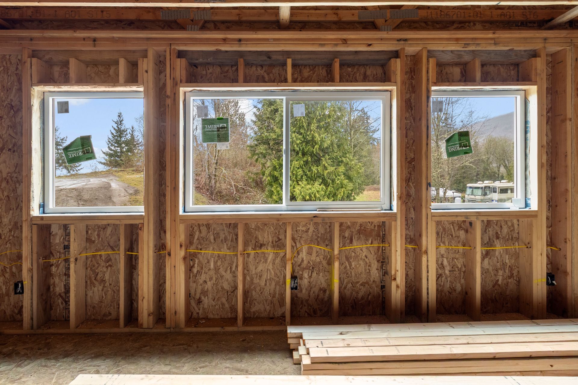 Three windows framed in unfinished wood studs inside a house under construction, overlooking a wooded landscape.