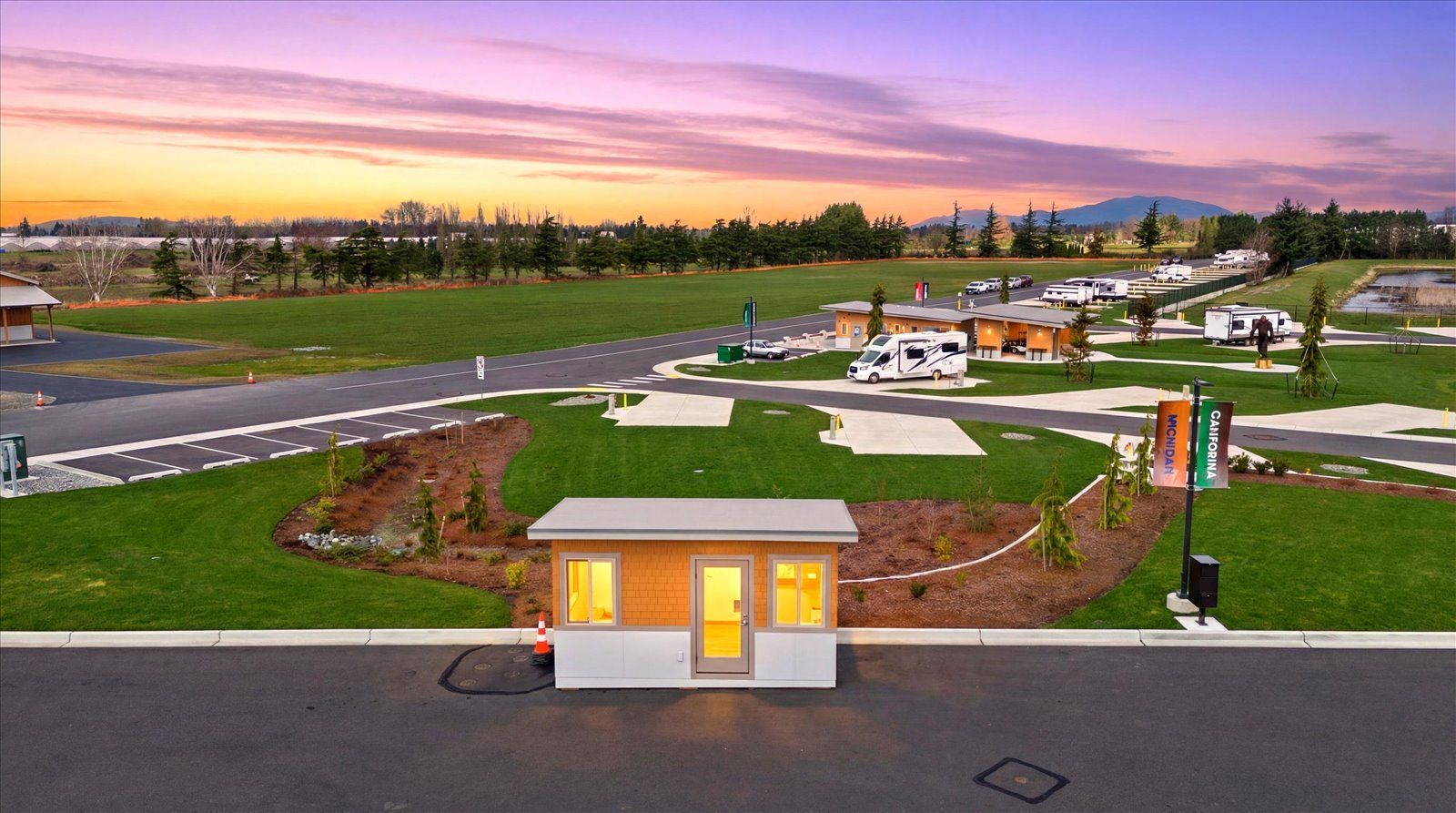 A small security booth sits in front of a modern RV park at sunset, featuring paved roads and several parked motorhomes.