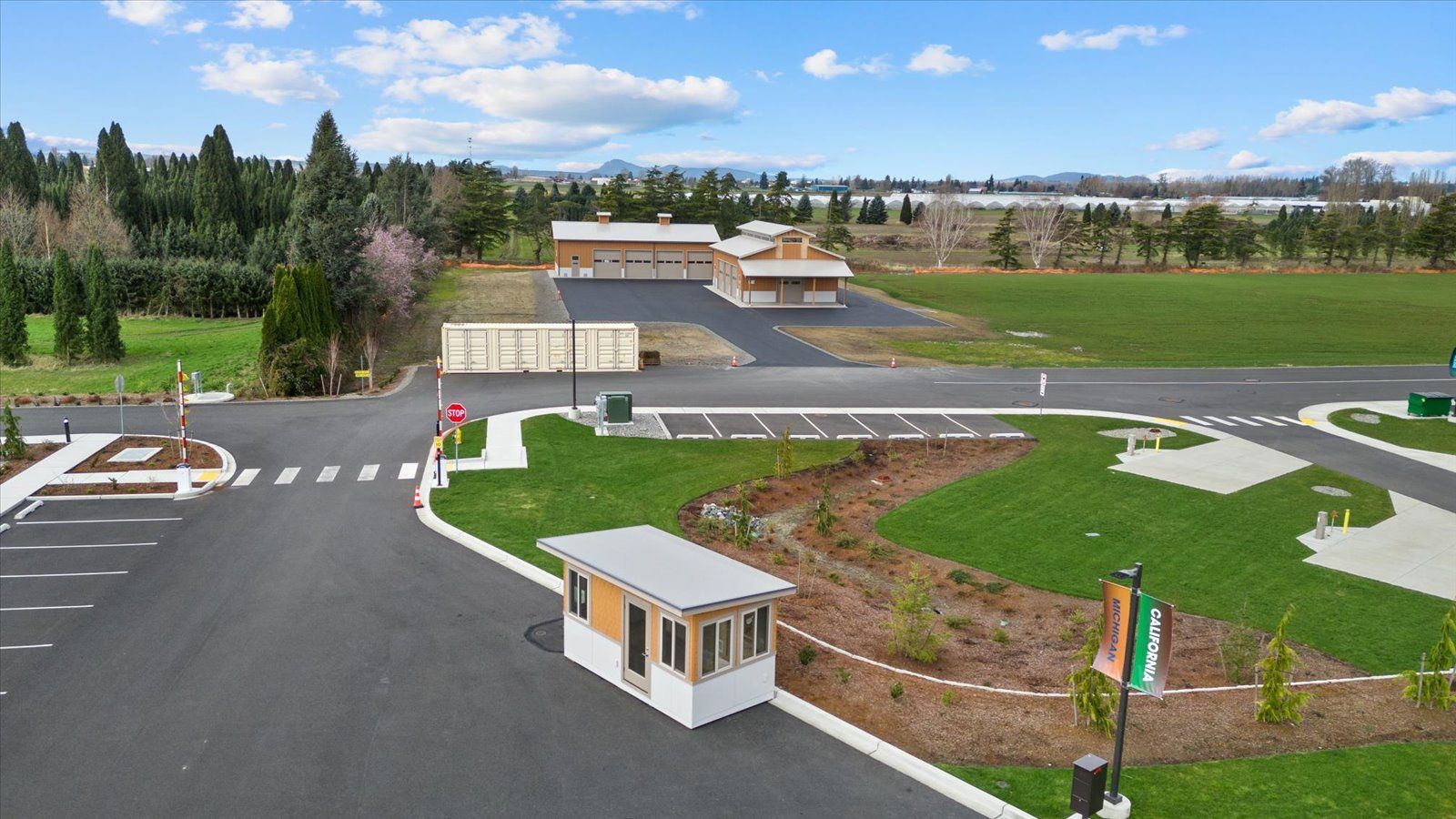 A small security booth stands at the entrance of a parking lot near a grassy field and a distant building.