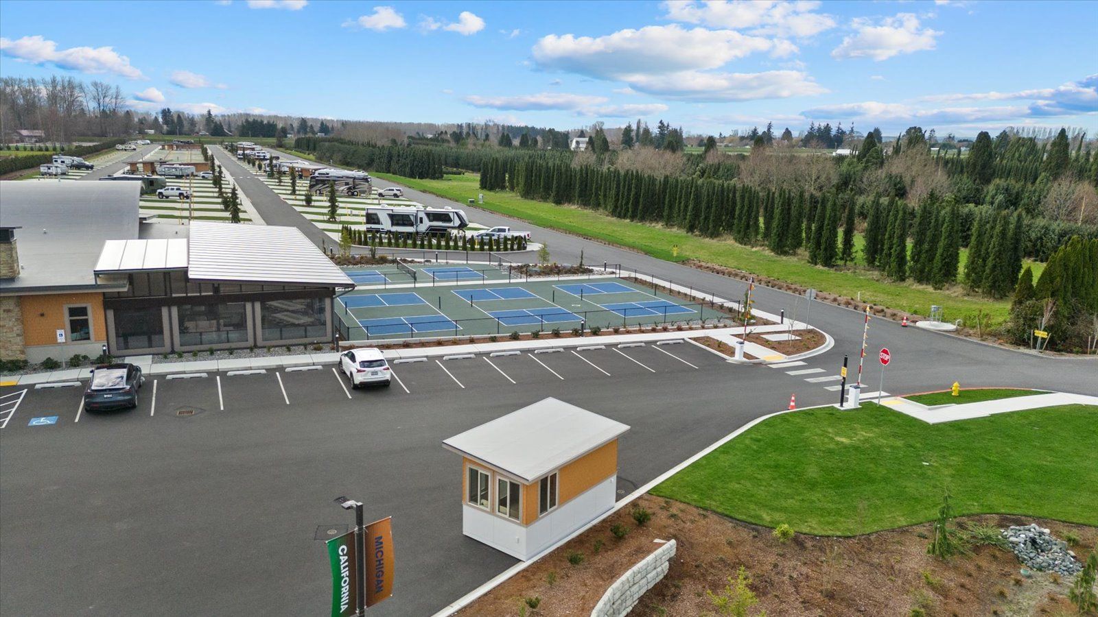 An aerial view of an RV resort with paved parking, tennis courts, a small booth, and rows of parked vehicles.