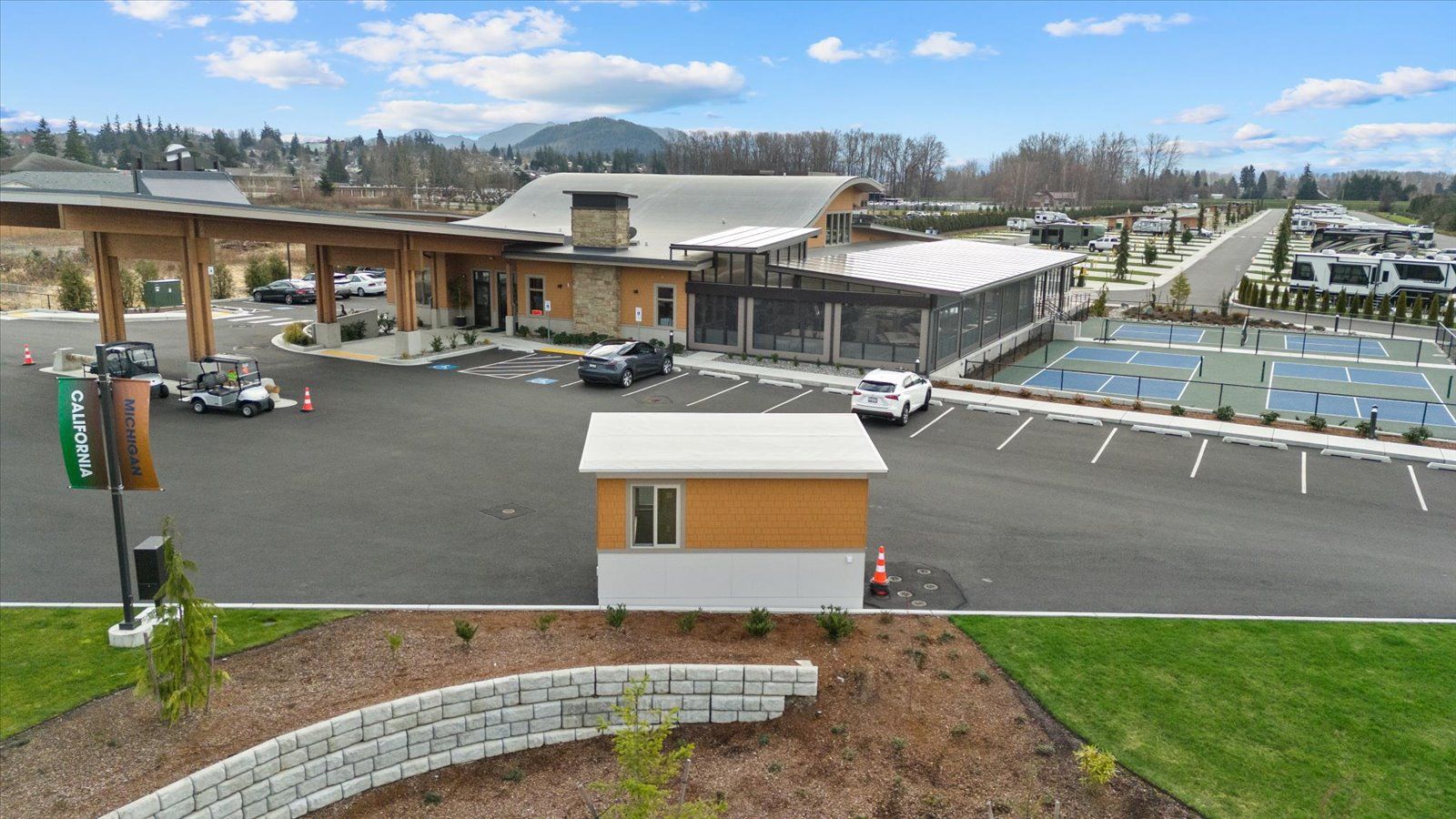 A modern resort building with a covered entrance, a small booth, a parking lot, and sports courts under a blue sky.