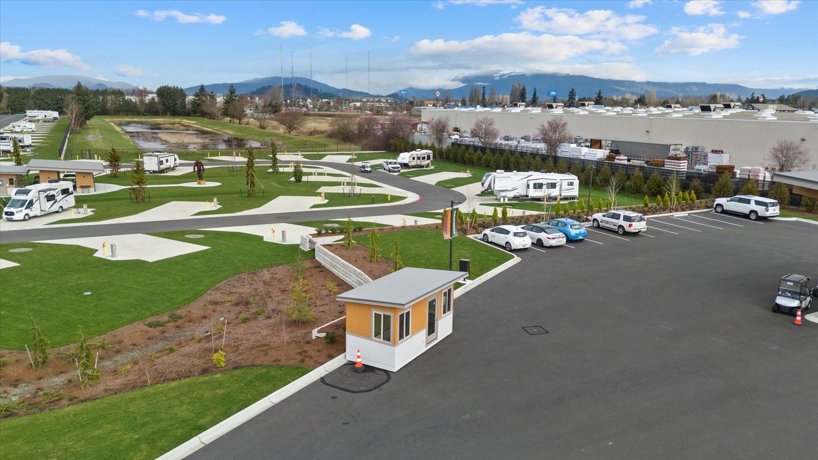 An aerial view of an RV park with several campers parked on green lawn spaces, a small office building, and a parking lot.