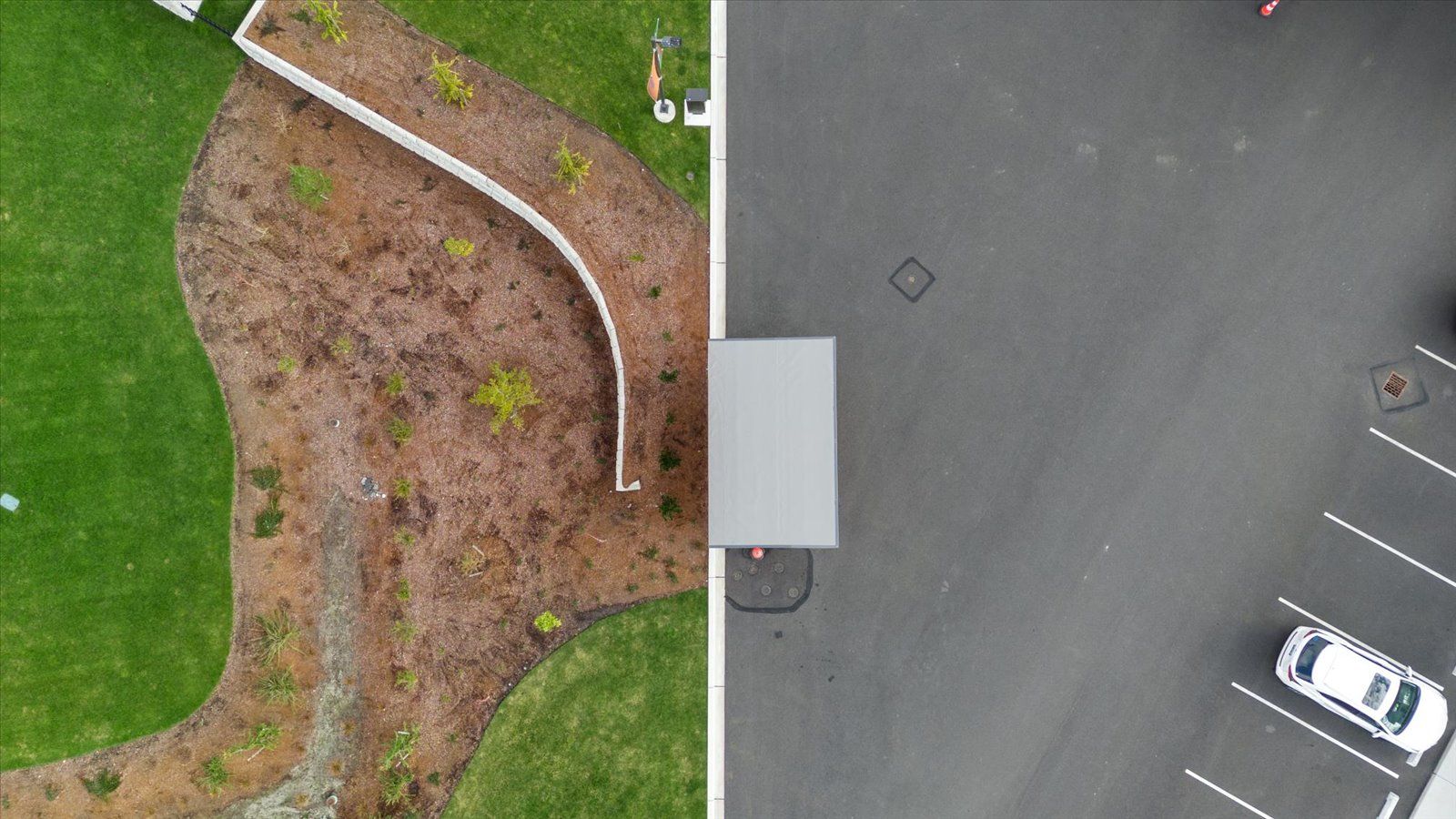 An aerial view of a landscape featuring a curved stone garden wall, lush green lawn, and an adjacent asphalt parking lot.