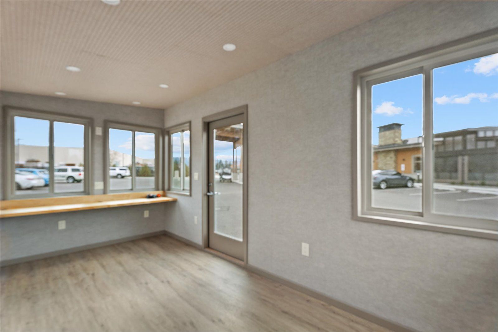 A vacant, modern room with light wood flooring, gray walls, recessed lighting, a wooden counter, and several windows.