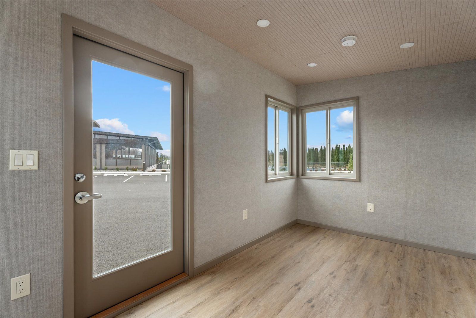 Empty room with grey textured walls, light wood flooring, a door, and two windows looking out onto a parking lot.