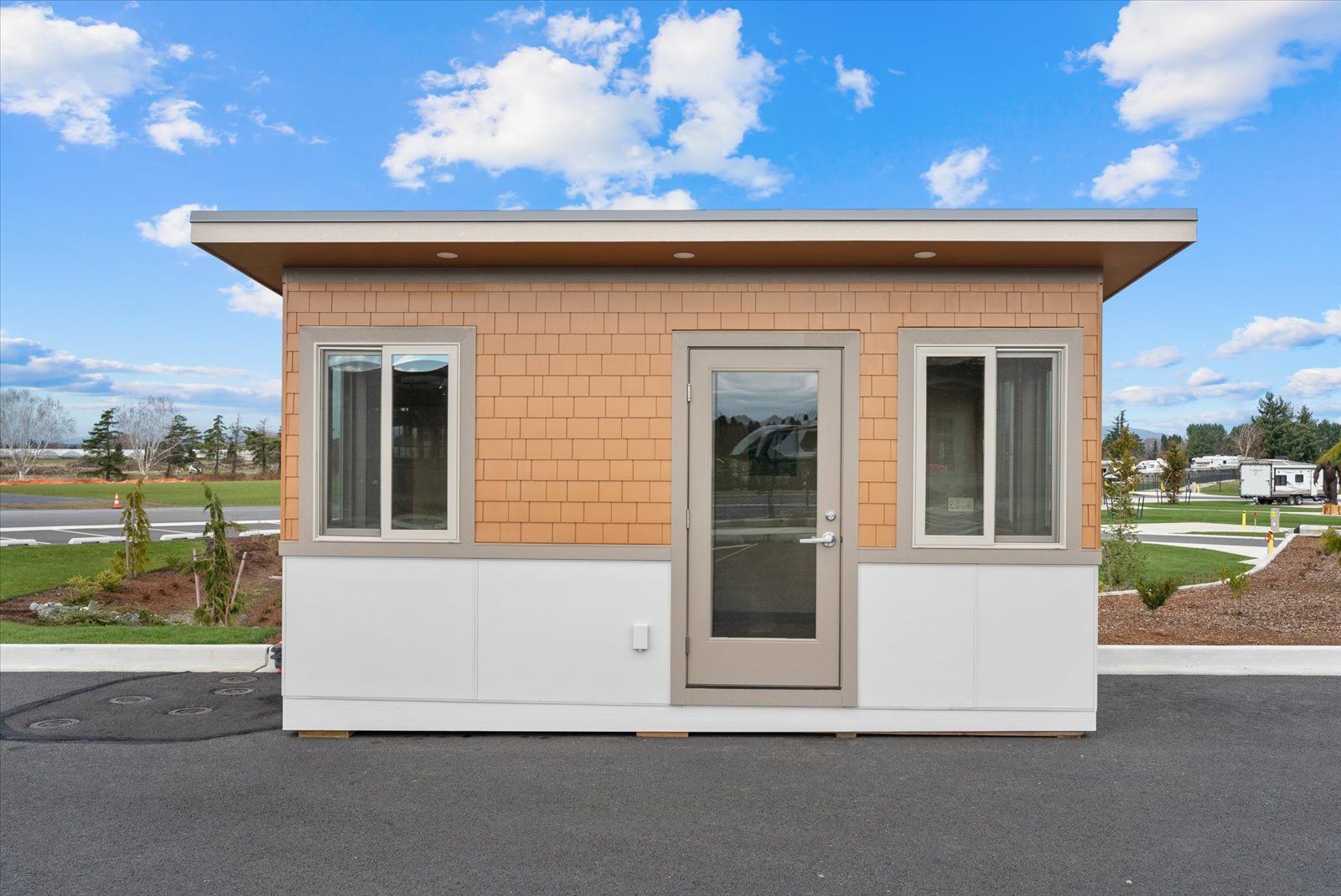 A small, single-story security or office booth with tan shingle siding, a central glass door, and two side windows.