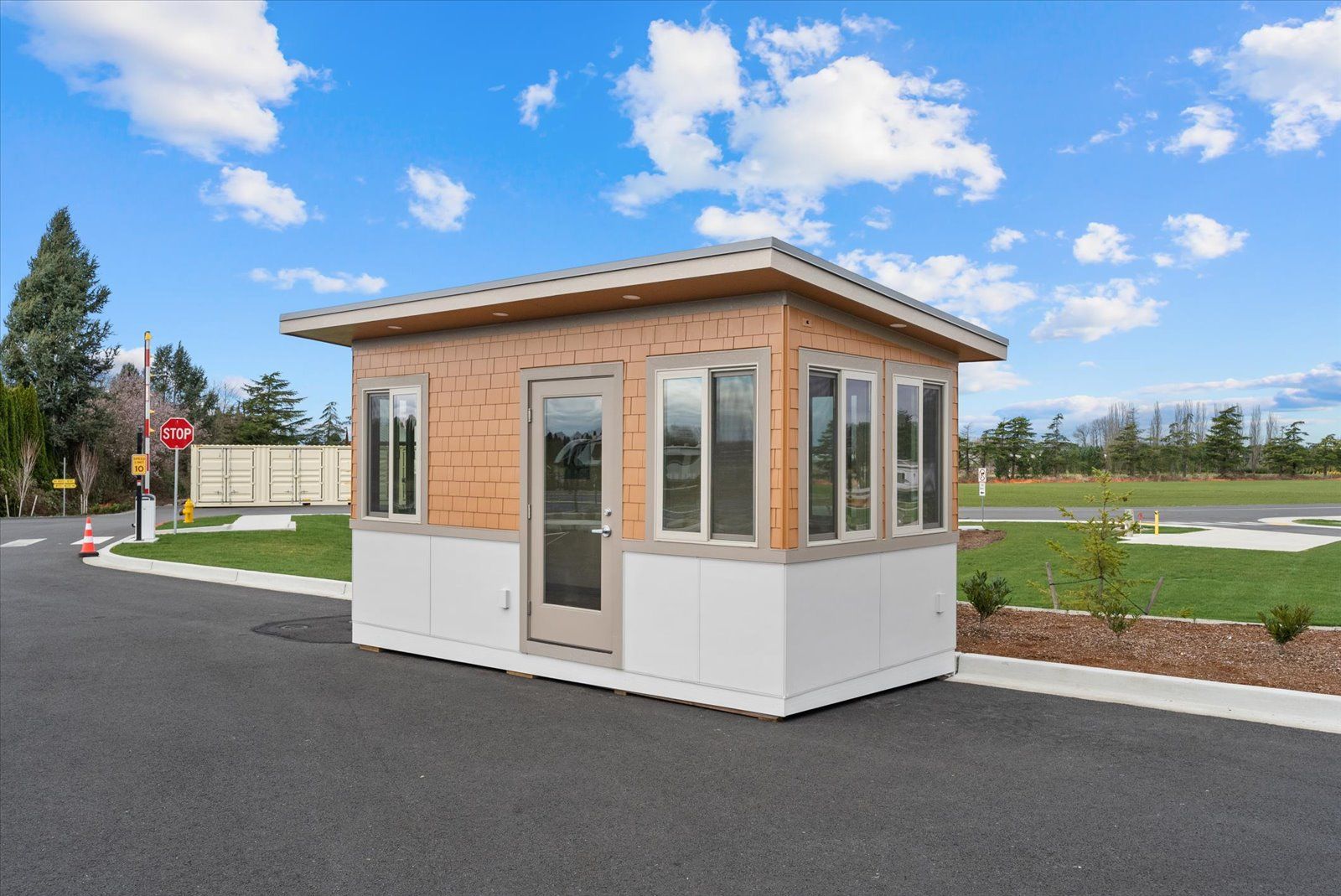 A modern security booth with a tan wooden upper exterior and white lower panels, situated on an asphalt lot.