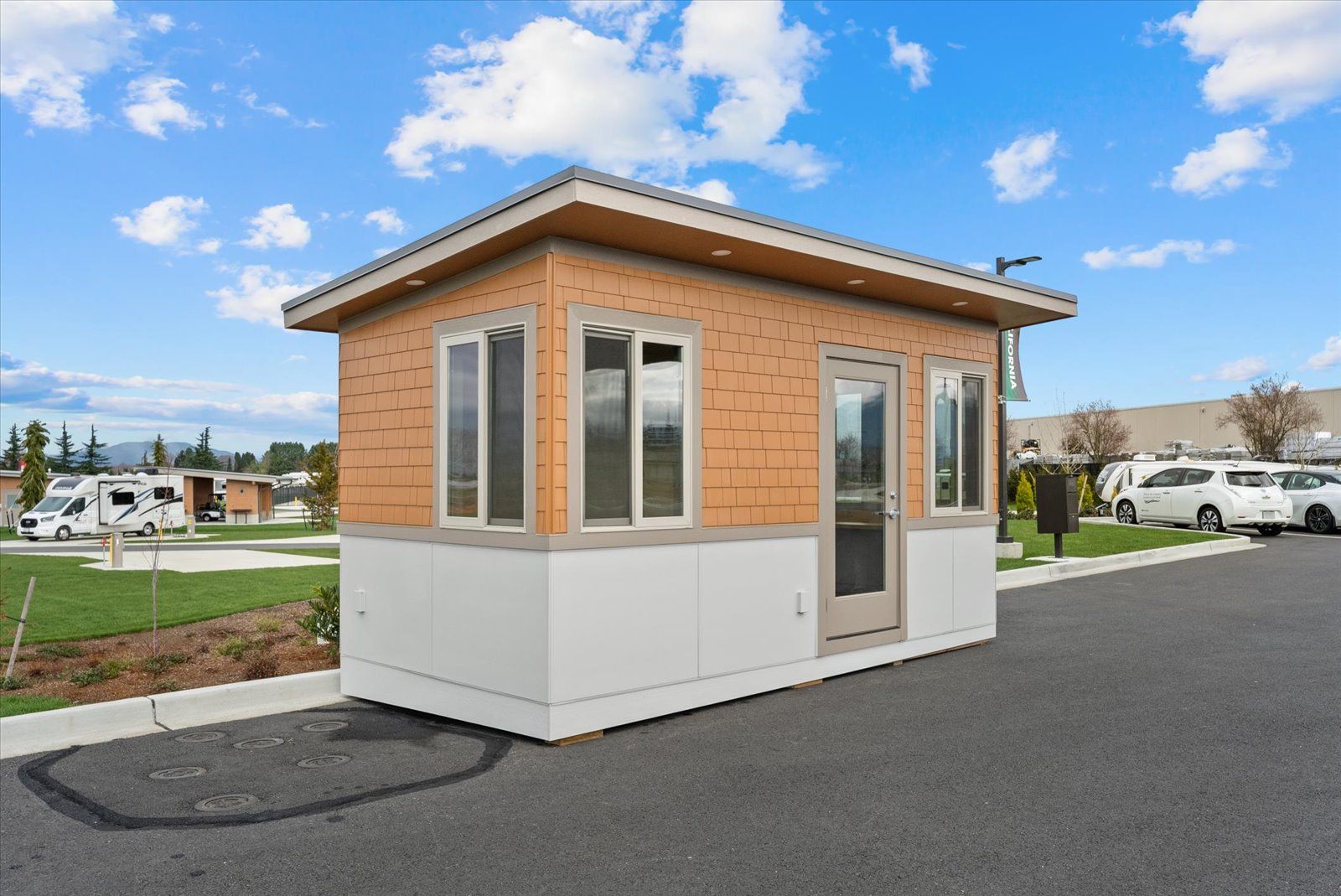 A small, tan and white rectangular guard booth sits on an asphalt lot near a green lawn and parked vehicles.