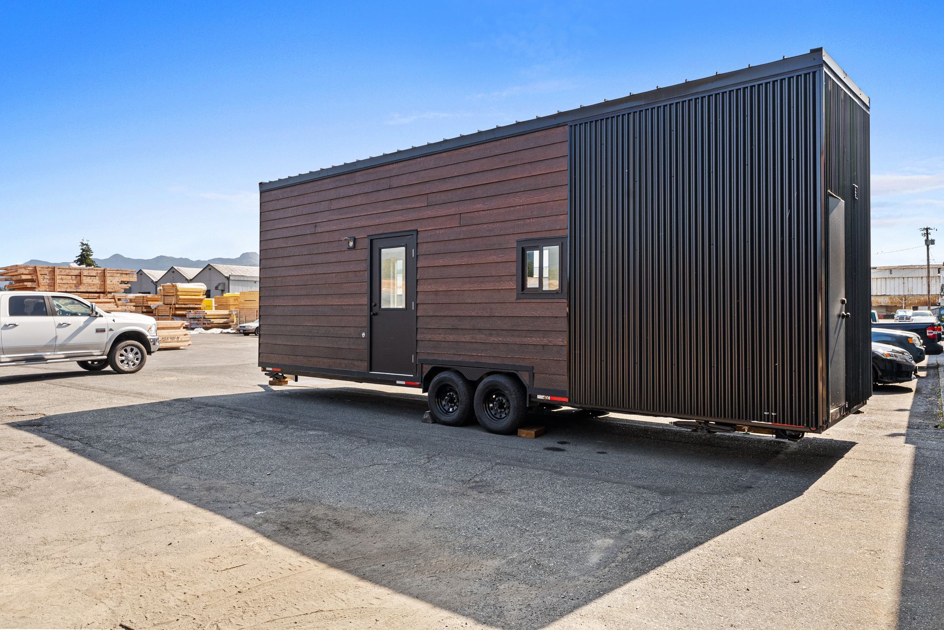 A tiny house on a trailer is parked in a gravel lot next to a truck.