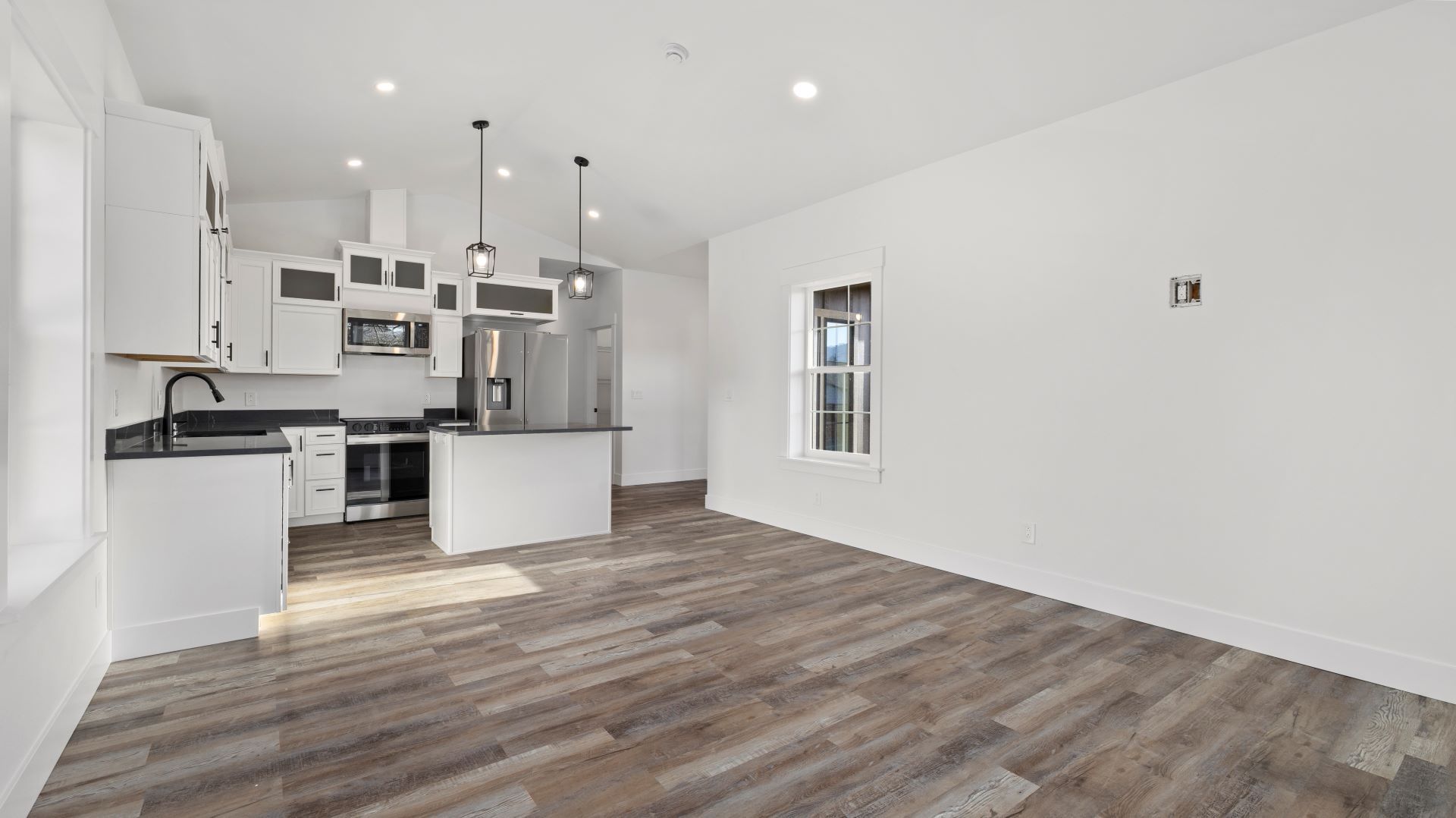 An empty living room with hardwood floors and a kitchen in the background.