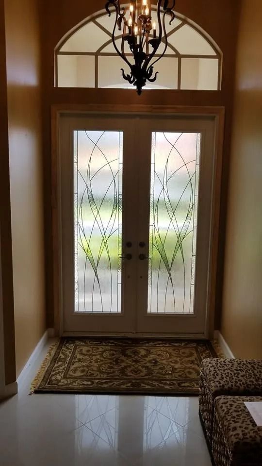 An entryway with white double doors featuring patterned glass, a dark arched transom window, a chandelier, and a floor mat.