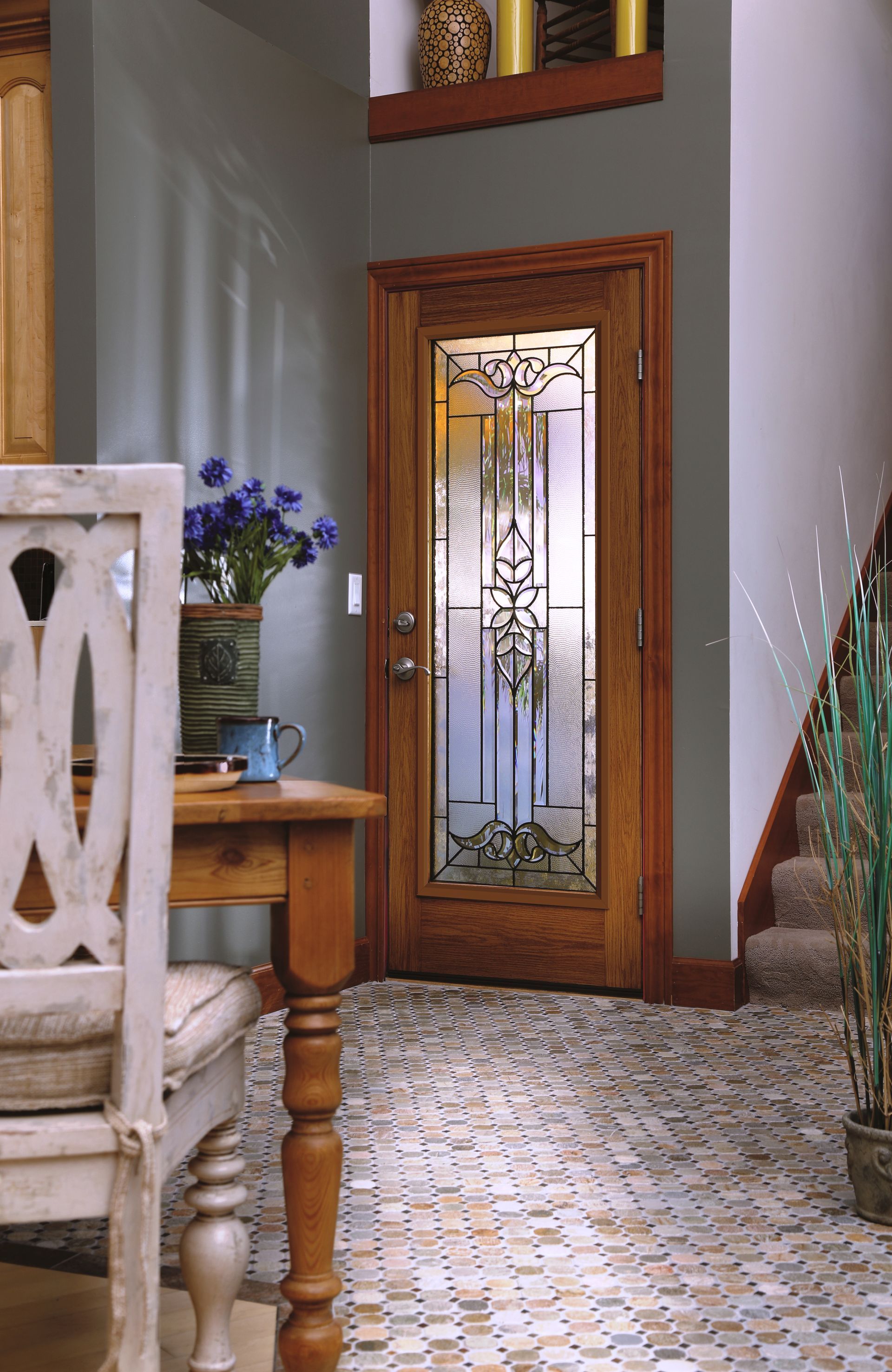 A wood front door with decorative stained glass in a home entryway with a tiled floor, dining chair, and grey walls.