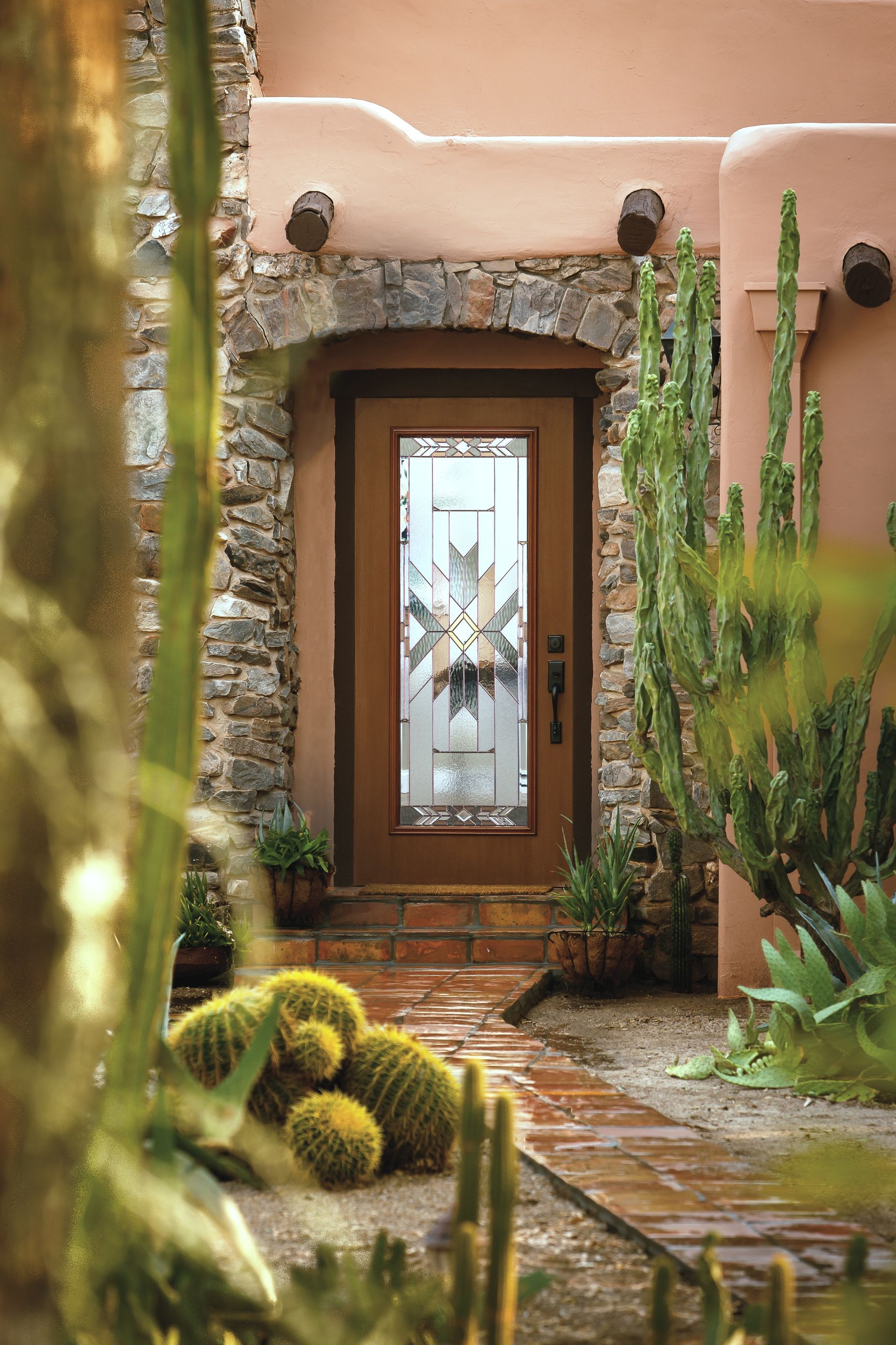 A front door with a decorative glass window, framed by stone and pink stucco walls, surrounded by desert cacti.