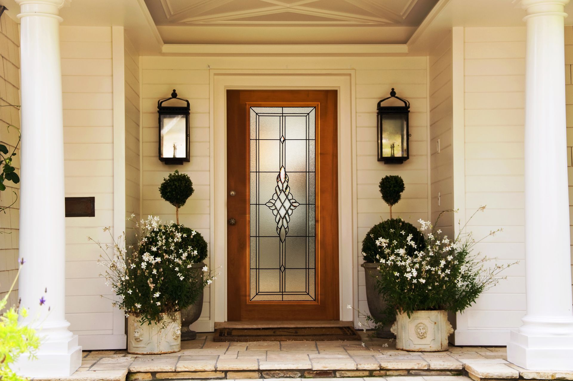 Front entrance with a wooden glass-paneled door, flanked by two white pillars, hanging lanterns, and potted green shrubs.
