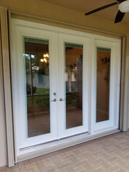 White french doors with glass panels on a brick patio. Brown trim surrounds the doors, and the ceiling has a ceiling fan.