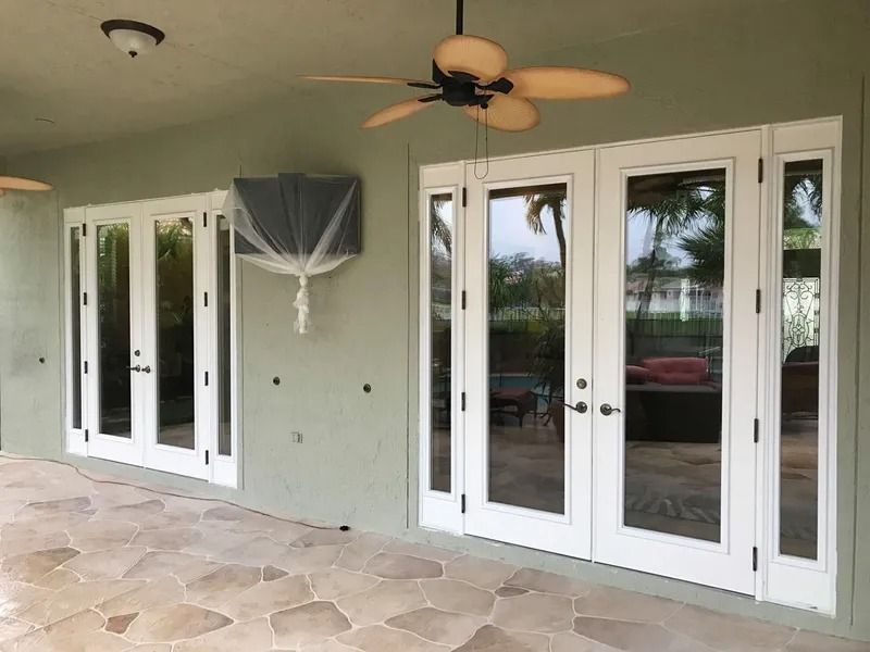White-framed French doors on a stone patio under a green roof, with a TV covered in plastic and a ceiling fan.