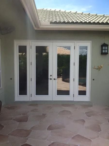 White French doors on a beige stone patio, flanked by light green stucco walls and a tiled roof, bright sunlight.