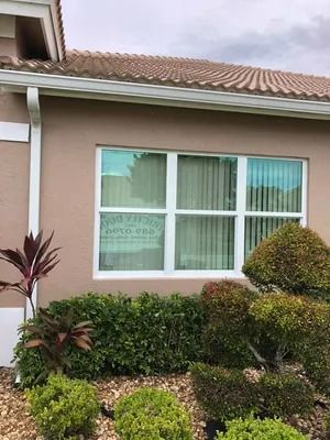 Tan house exterior with white-framed window. Green bushes and red plant in front. Window has vertical blinds.