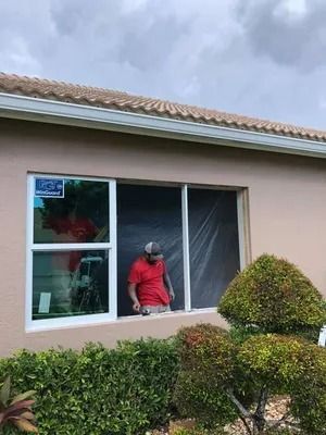 Man installing window in beige house with green bushes under cloudy sky.