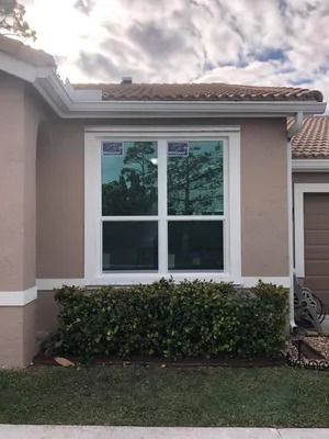 White-framed window on a tan house with green bushes and grass in front. Cloudy sky overhead.
