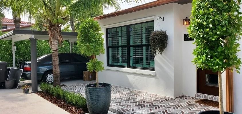A white stucco house with black shutters, a driveway, and potted trees. A car is parked under a carport.