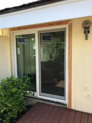 A newly installed sliding glass door with wood trim, on a cream-colored house, next to a bush and wooden deck.