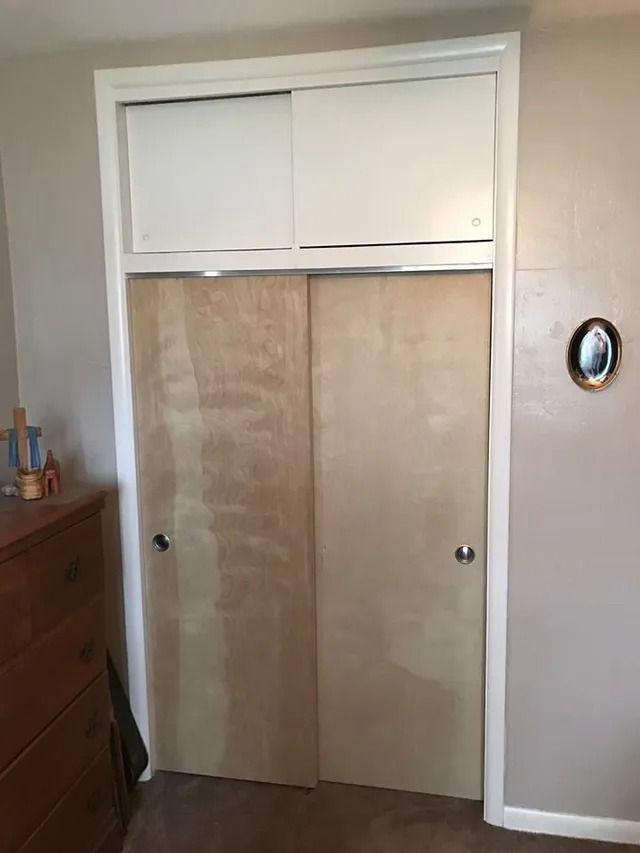 A closed closet with light-colored plywood doors, white trim, and a white sliding panel above.