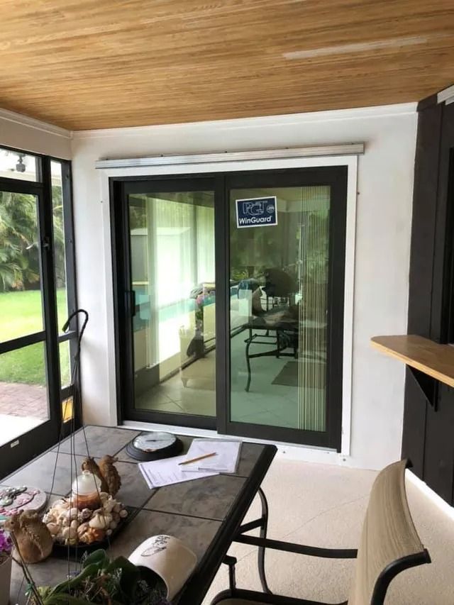 Sliding glass door inside a screened porch. Black frame, white trim, wood ceiling. Table with objects visible.