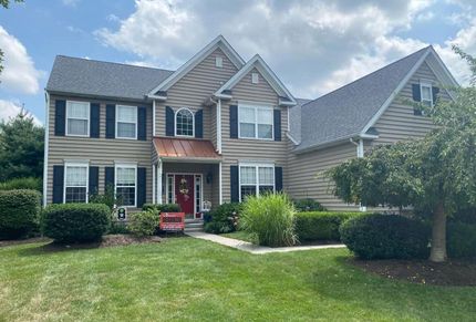 Two-story beige house with black shutters, red front door, and a green lawn under a cloudy sky