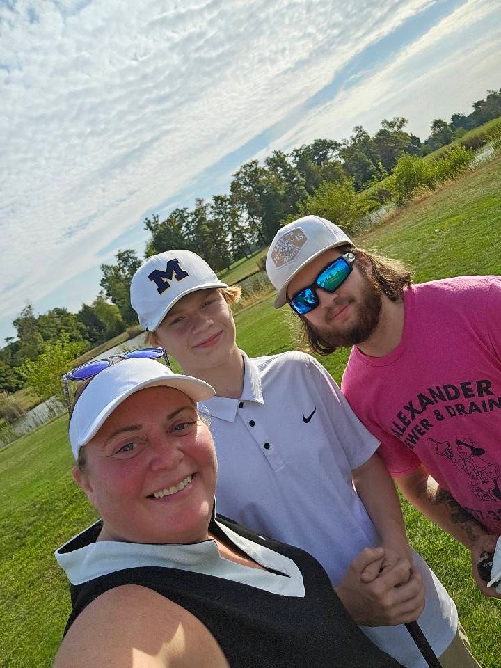 Group of three people smiling on a golf course under a cloudy sky. One woman and two men, all in golf attire.