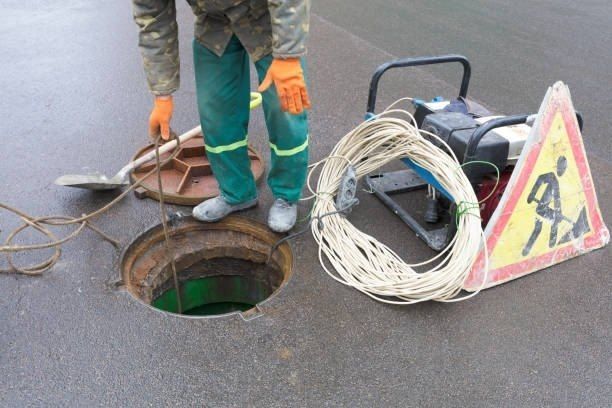 Manhole open on wet pavement, worker with orange gloves, caution sign, generator, and cable.