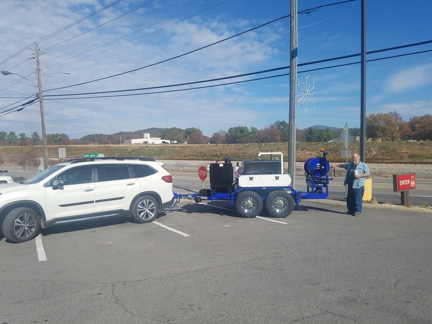 White SUV towing a blue trailer with equipment, parked near railroad tracks; person standing nearby.