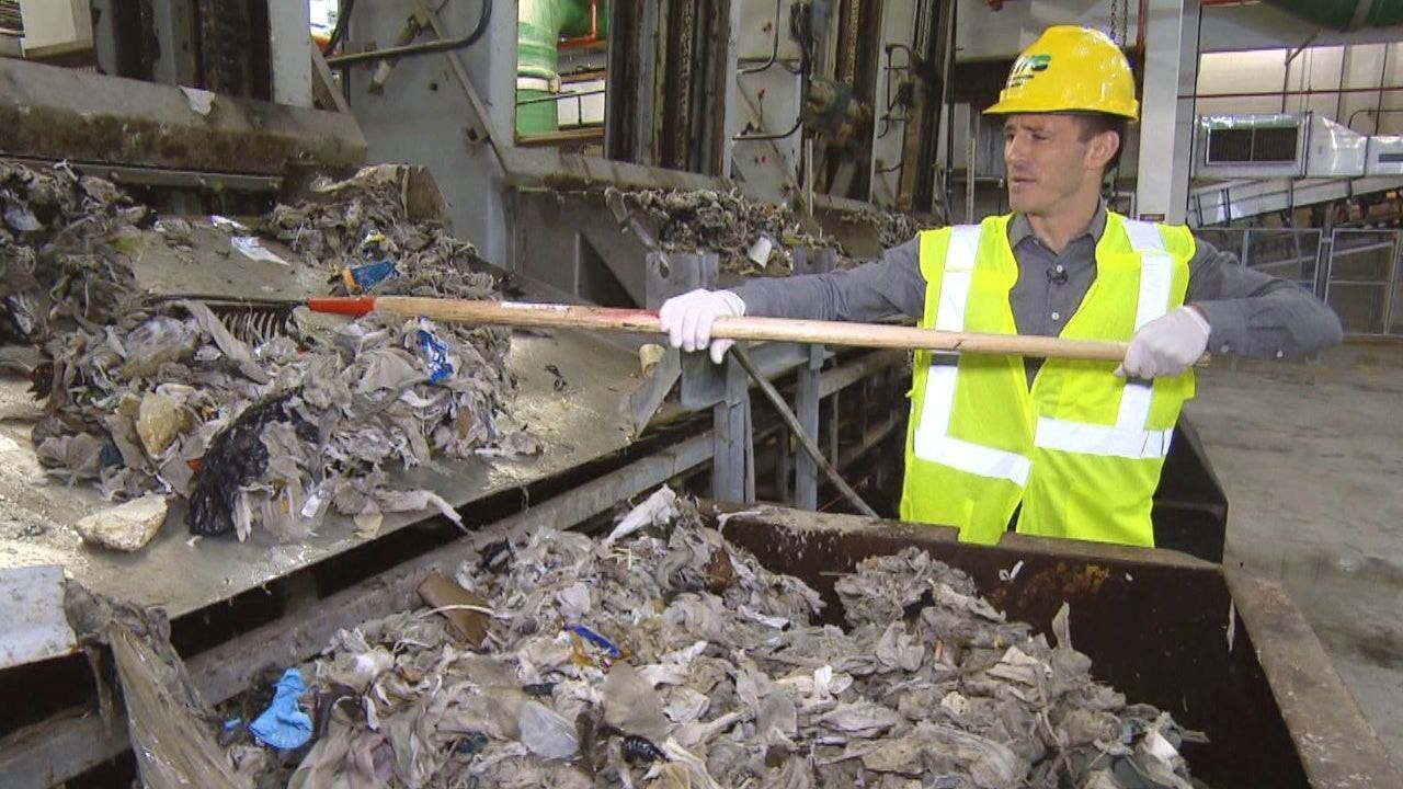 Man in safety gear sorts trash with a shovel in an industrial waste processing facility.