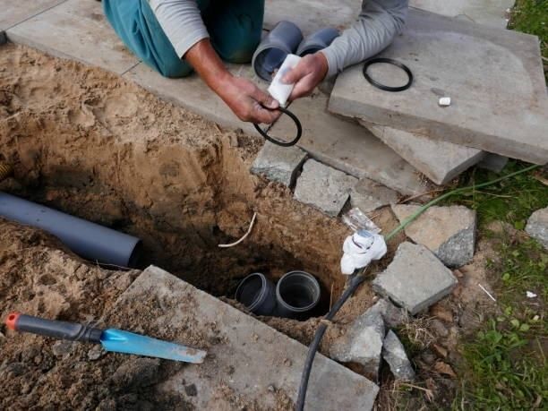 Person installing plumbing pipes in a trench, using gaskets and other tools outdoors.