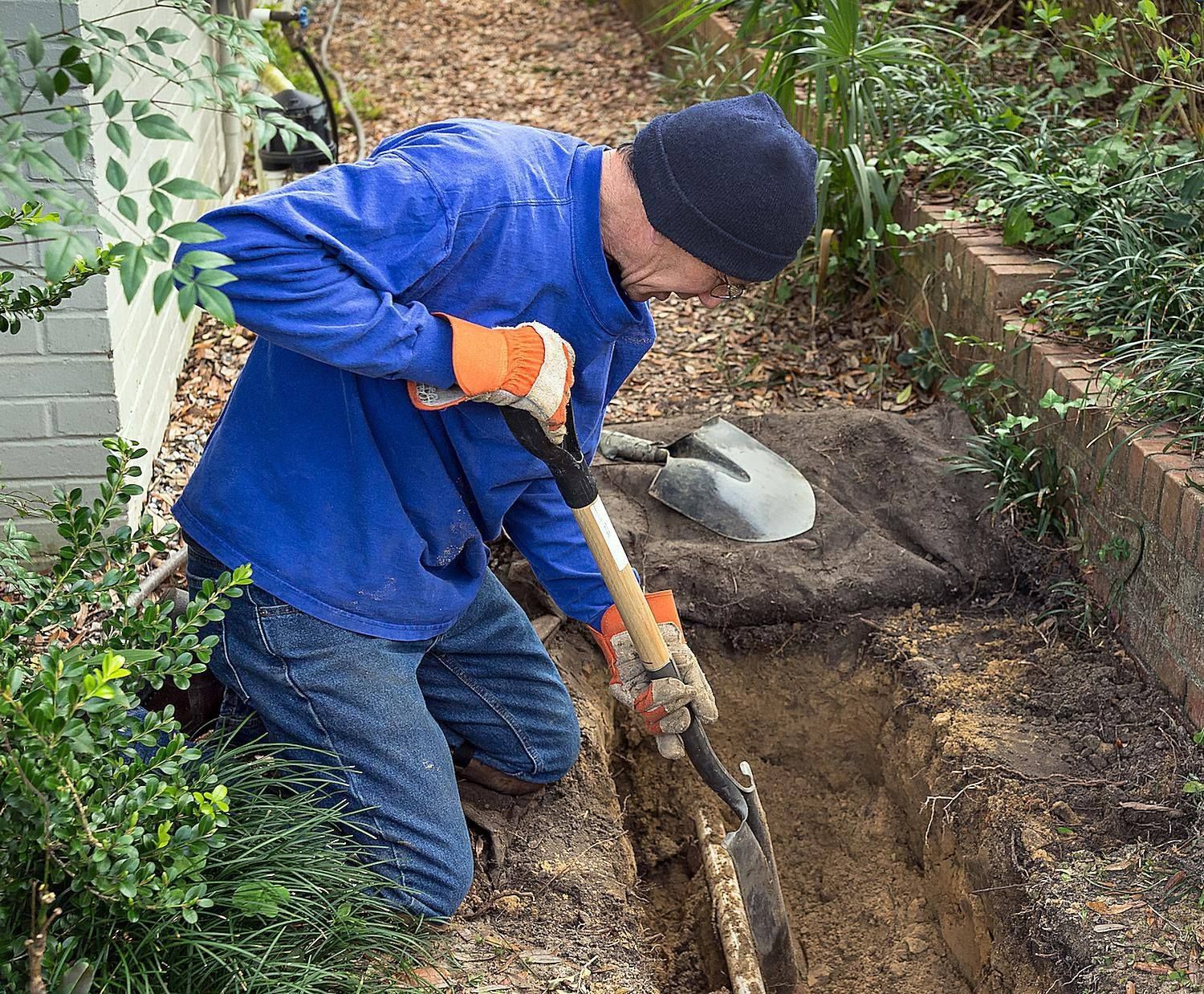 Man kneeling, digging a trench in a garden bed, wearing blue shirt and orange gloves.