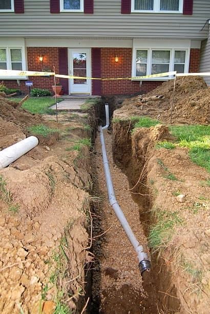 Trench dug in front yard of house with installed pipe for drainage, yellow caution tape visible.