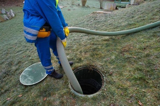 Person in blue overalls pumping a septic tank with a hose in a grassy yard.