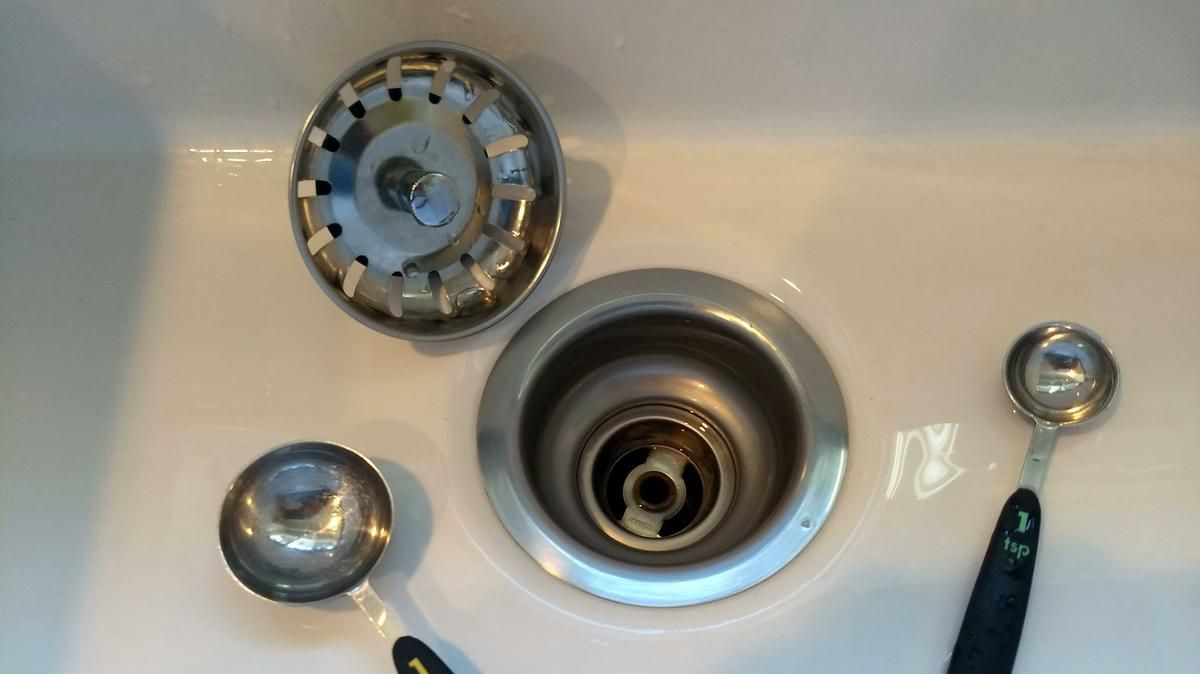 Sink drain components laid out in a white sink. Includes a strainer, drain, and measuring spoons.